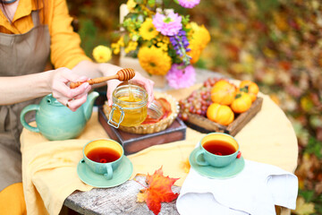 Autumn picnic. Woman in yellow dress and linen apron drinks tea from cup at wooden table in garden. Beautiful kettle, tablecloth, honey with spoon, apple pie, harvest, persimmon, grapes, maple leaf