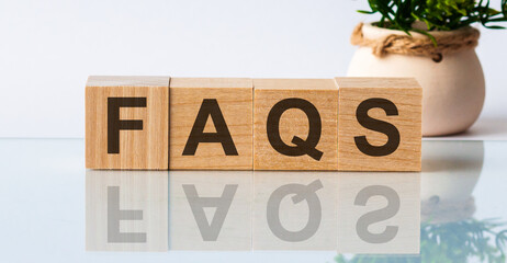 FAQS is a word written in black letters on wooden cubes located on a white mirror surface. The inscription is reflected, in the background there is a flower in a pot of light brown color.