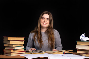 young girl sits at a table with books and documents against a black wall. Remote education. the teacher is preparing for the lesson. office worker in a working environment. look at the camera