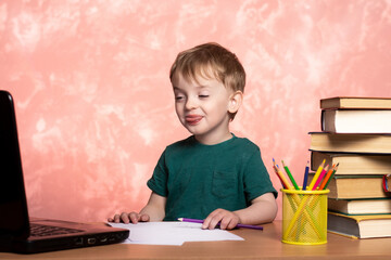 little boy sitting at home at the table and shows tongue in a laptop. the difficulties of quarantined distance education. a lot of books and pencils nearby. the child does not want to do homework.