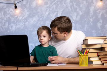 dad and son are sitting at home at the table with books and watching online lessons on a laptop. quarantined distance learning. home family leisure and comfort.