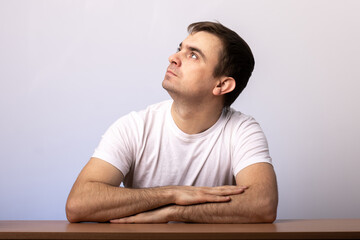 young male student sitting at a desk with folded arms. pensive look up. White T-shirt. Gray background. place for text. office employee at work.