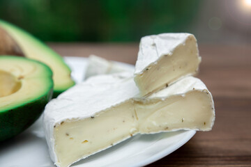 sliced circle of brie cheese or camembert with mold on a white plate. cut avocado on the background in defocus. healthy diet.
