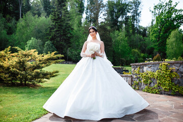 Fototapeta premium Young beautiful bride is standing on a garden. Beautiful bride in white wedding dress with bouquet in her hands the wind blows the veil. Sunshine portrait of happy bride outdoor in nature.