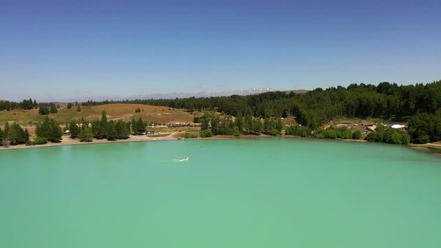 Aerial tilt down shot of turquoise lake near forest against sky, drone flying forward towards person wakeboarding on sunny day - Lake Tekapo, New Zealand
