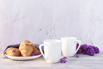 Two beautiful porcelain cups of coffee with milk with croissants decorated with lilac flowers on white wooden table. Perfect breakfast concept. Copy space.