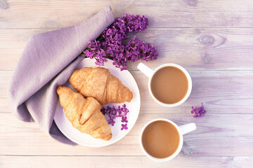 Two beautiful porcelain cups of coffee with milk with croissants decorated with lilac flowers on white wooden table. Perfect breakfast concept. Flat lay style.