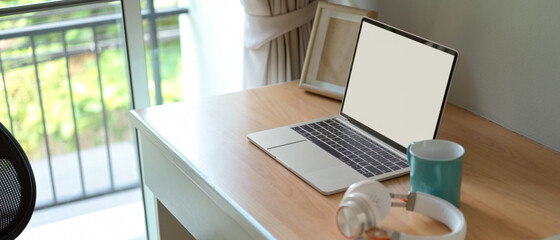 Simple worktable with blank screen laptop, mock-up frame, headphone and mug on wooden table in bed room