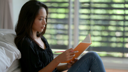 Fototapeta premium Female freelancer looking on her schedule book while sitting on floor at bedroom