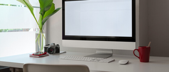 Stylish office desk with blank screen computer, camera, mug, office supplies and decoration
