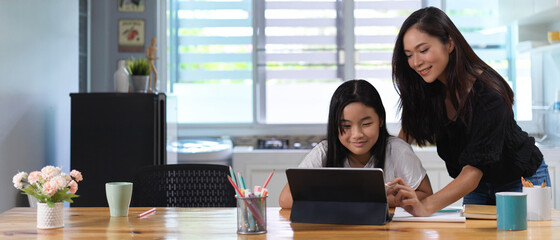 Mother helping her daughter do homework with digital tablet on wooden worktable in living room
