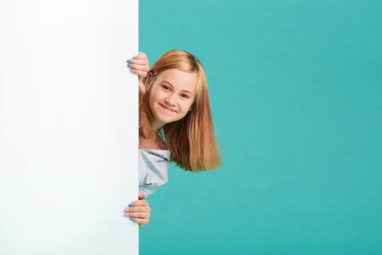 A smiling girl stands behind a white blank panel isolated against a turquoise background. Looks out from behind the banner, an empty space for the text.