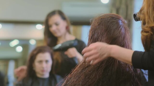 woman master dries brunette long hair with hairdryer reflecting in large beauty salon mirror slow motion
