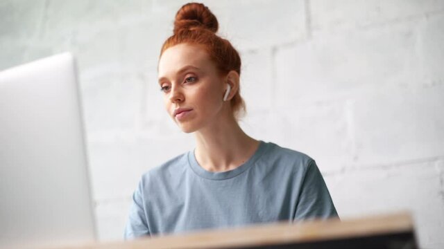 Close-up of face of beautiful redhead young woman working on laptop and filling out data in paper documents, shooting from below. Girl manager working in home office and listening music in headphones.