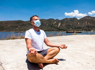 Coronavirus COVID 19. Young beautiful man doing sport performing workouts exercises yoga in the park near sea during quarantine. Active life in surgical sterilizing face mask protection