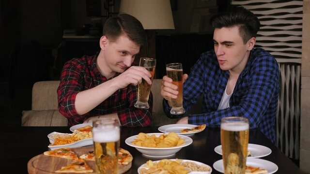 Two men drink beer in a cafe, they clink glasses and talk. Friends relax in a pizzeria, drink beer, eat pizza, potato chips and snacks. Slow motion.