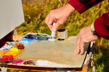 Male hand of artist, palette with paints and canvas in nature outdoors and greenery background