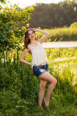 Naklejka na ściany i meble Beautiful smiling girl in a white shirt and denim shorts standing near the river