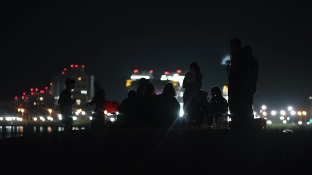 Young people sitting on the beach at night on the background of glowing skyscrapers and having fun - smoking hookah and dancing