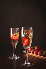 Two glasses of cold splashing champagne against wood plate with strawberries and dark background