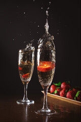 Two glasses of cold splashing champagne against wood plate with strawberries and dark background