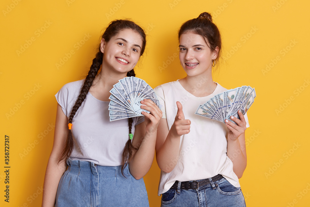 Wall mural two friends holding cash in hands, looking at camera with happy facial expression, ladies with dolla