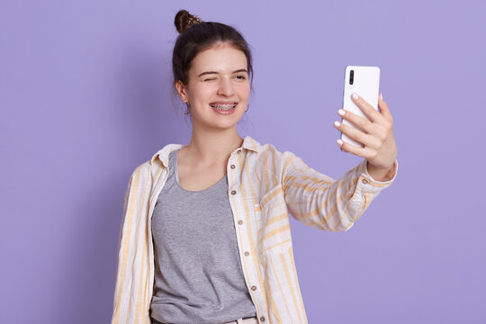 Happy funny woman posing isolated over lilac background, winks eye and taking selfie, woman with dark hair and bun standing isolated over lilac background.