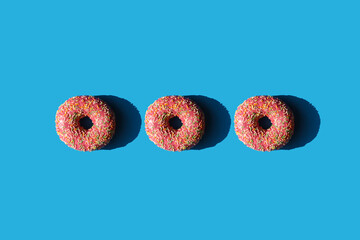 Three pink donuts in a row on a blue background with hard shadows flat lay