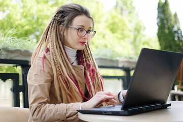 Concentrated female journalist in eyeglasses for vision correction and long dreadlocks hair is writing a questions for next interview using laptop computer while sitting in coffee shop outdoors.