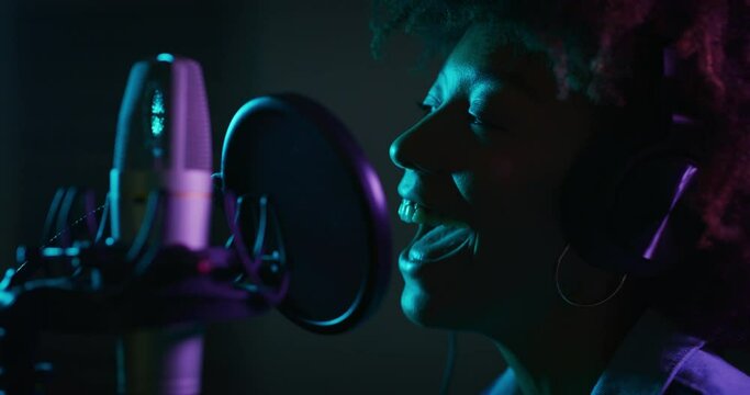 An young professional smiling energetic african female singer wearing headphones is performing a new song with a microphone while recording it in a music studio with colorful lights on a background.