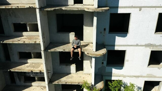 Male athlete showing muscles outside derelict building, Zoom out drone view of strong sportsman demonstrating biceps while sitting on balcony of desolate building during break in workout on summer day