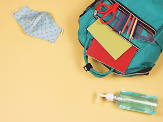 COVID-19 prevention while going back  to school  and new normal  concept. Top view of backpack with school supplies , blue polka dot fabric mask and alcohol sanitizer gel on yellow background