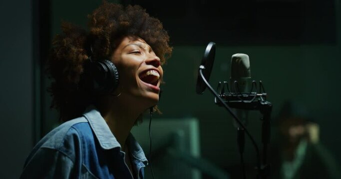 An young professional african female singer is performing a new song with a microphone while recording it in a music studio. 
