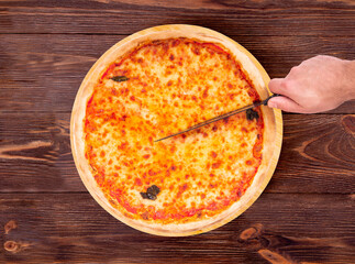 Top view of male hand which cutting the pizza with a kitchen knife. Delicious pizza with mozzarella, bocconcini and basil leaves or pizza margherita on a wood platter which is on wooden rustic table