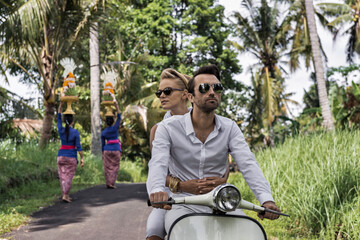 Asia, Indonesia, Bali, young Caucasian couple, wearing smart casual clothing, enjoying a ride on a classic  scooter through typical Balinese scenery, passing local women carrying temple offerings.