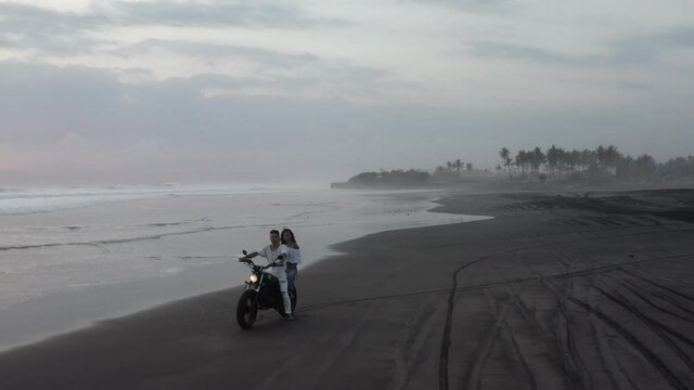 aerial of couple on motorcycle at black sand pasut beach in Bali Indonesia during sunset