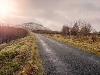 The Gleniff Horseshoe loop drive in county Sligo, Ireland, Winter season. Mountains covered with snow. Nobody, Cloudy sky. Small country road lead into mountains, Irish nature. Sun flare.