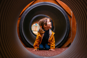 A little girl climbs in the pipes. Modern and safe children's Playground. Round and long tunnel. Overcome obstacles. Children's curiosity and discovery of everything new.
