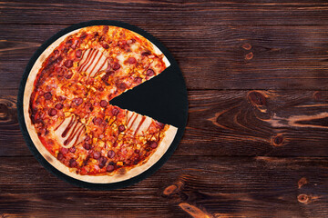 BBQ pizza with ham, bbq sauce, bacon and salami, without one slice on a slate black platter which is on wooden background, top view and copy space