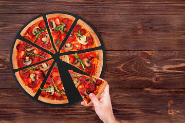 Fototapeta na wymiar Young male taking a slice of tasty pizza hot with salami, arugula, cherry tomatoes, mushrooms and texas spice mix, from the slate black platter which is on wooden rustic table