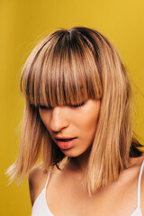 Side view of stylish young woman with trendy modern haircut posing on camera. Model looks down showing her blonde hair with bangs. Isolated over yellow background.