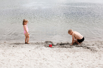 Brother and sister are playing on the lake, on a sandy beach near the water. A seven-year-old boy and a three-year-old girl in a swimsuit build sand castles. Children's friendship. Happy summer holida