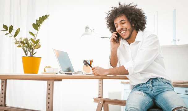 Side view of young entrepreneur or student talking mobile phone while working from home. Charming Arab guy chatting on smart phone with friends or customers. Tinted image.