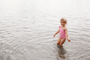 A three-year-old girl of European appearance plays in the water on the beach. A child walks ankle-deep in the lake. Holidays in nature. Summer photo of a baby in a swimsuit.