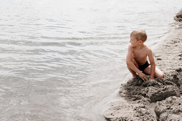 Fototapeta premium Seven-year-old boy of European appearance plays with sand on the beach. A child sits on the seashore. Happy vacation in nature. 