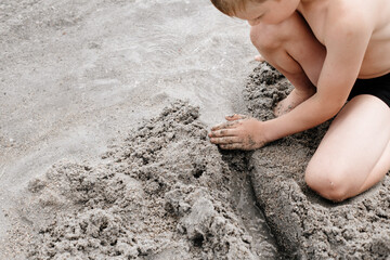 Seven-year-old boy of European appearance plays with sand on the beach. A child sits on the seashore. Happy vacation in nature.
