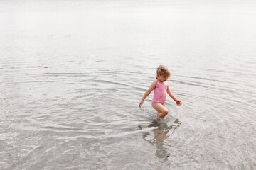 A three-year-old girl of European appearance plays in the water on the beach. A child walks ankle-deep in the lake. Holidays in nature. Summer photo of a baby in a swimsuit.