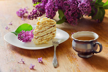 .A piece of cake lies on a plate with lilac flowers on the table and a mug of tea. Breakfast