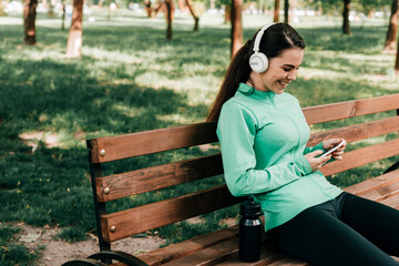 Smiling sportswoman in headphones using smartphone near sports bottle on bench in park