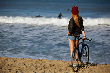 Attractive hipster girl relaxing on the beach after ride on her sport fixed gear bicycle, young woman standing on seashore looking to surfing people in water, charming female with rental bike outdoors